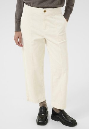Pantaloni in corduroy crema con design a gamba dritta, dotati di chiusura anteriore con bottone e due tasche laterali. Indossati con scarpe nere e calzini marroni.