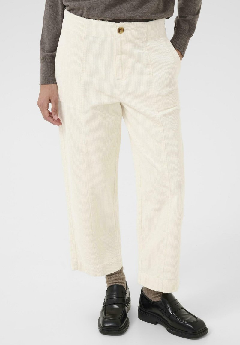 Pantalon en velours côtelé crème avec une coupe droite, présentant une fermeture à bouton à l'avant et deux poches latérales. Porté avec des chaussures noires et des chaussettes marron.