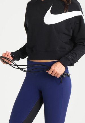 Kobieta ubrana w czarną bluzę Nike i niebieskie legginsy trzyma czarną skakankę na wysokości talii, na jednolitym tle.