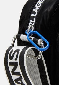 Podrobnosti o črni najlonasti torbi s plavim karabinom, črno-belim trakastim ročajem in bold logotipom. Gladka tekstura, kompaktna zasnova.