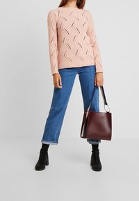 Lichtroze gebreide trui met golvende textuurpatronen, gecombineerd met blauwe wijde jeans en zwarte haklaarzen. Houdt een bordeauxrode emmer tas vast.