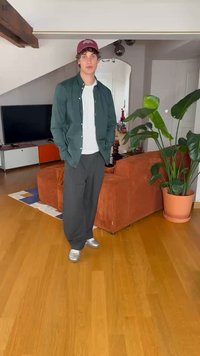 Grøn skjorte med knapper over en hvid t-shirt, sorte brede bukser og sølvfarvede sneakers. Står i et rum med en plante og en orange sofa.