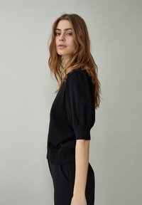 Jonge vrouw met lang bruin haar, gekleed in een zwart kortermig shirt en een zwarte broek, zijwaarts staand tegen een effen grijze achtergrond.