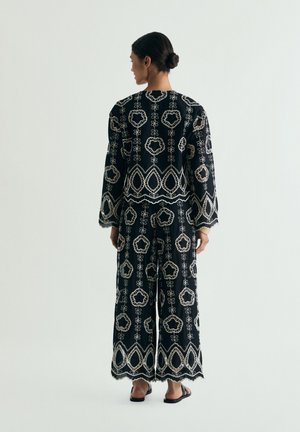 Donna con capelli scuri raccolti in uno chignon, indossa una maglia nera a maniche lunghe e pantaloni a gamba larga con ricami floreali bianchi, in piedi di spalle.