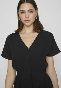 Čierne šaty s krátkym rukávom, so V-výstrihom, gombíkovou prednou časťou, nazhromaždeným pásom a splývavou sukňou. Hladká látka s jemnou textúrou.