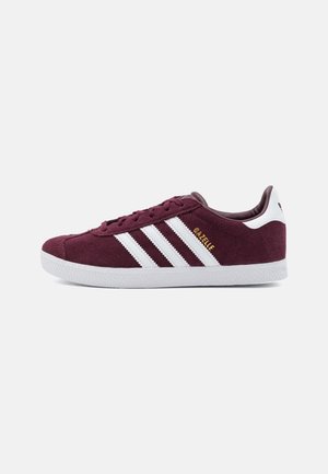 Baskets Adidas Gazelle bordeaux avec des bandes et une semelle blanches, inscription dorée "GAZELLE" sur le côté, vue latérale sur un fond uni.