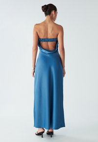 Modra satenasta maksi obleka z nabrano naramnico na hrbtu, ki ima izrezan detajl. Gladka tekstura in tekoč silhueta. Črne sandale na peto.