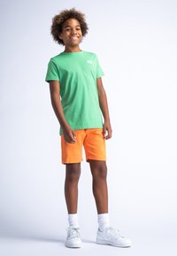 Groene katoenen t-shirt gecombineerd met oranje shorts. Opmerkelijke print op de shorts. Model draagt witte sneakers en sokken. Eenvoudige, casual outfit.