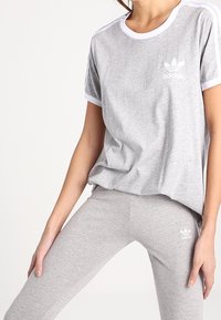 Šedé bavlněné tričko s bílým lemem a logem Adidas na prsou, doplněné o šedé legíny se shodným designem, které mají logo na lýtku.