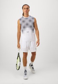 Top branco sem mangas com um padrão de pontos pretos em degradê, combinado com calções brancos. Modelo segura uma raquete de ténis, vestindo sapatilhas desportivas.