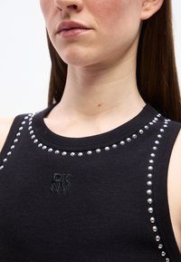 Κοντινό πλάνο γυναίκας που φοράει μαύρο αμάνικο τοπ με ασημένιες τρουκς που περιγράφουν το ντεκολτέ και τους ώμους, με λογότυπο DKNY.