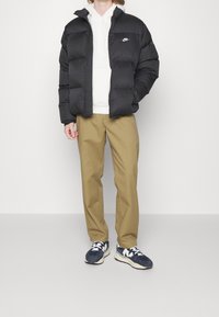 Persona vestida con una chaqueta de plumas negra de Nike, sudadera blanca, pantalones beige y zapatillas azules, de pie contra un fondo blanco liso.