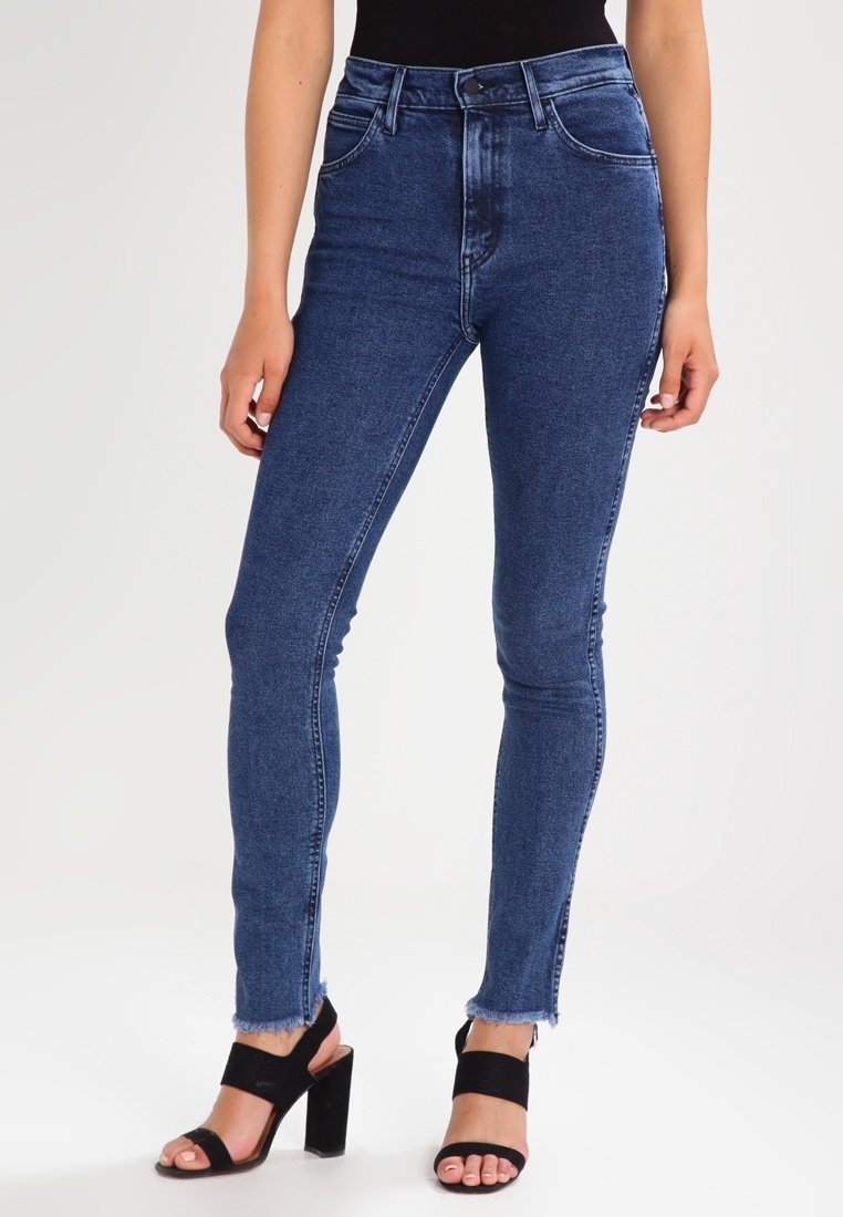 Jeans de mezclilla de cintura alta en azul oscuro con una silueta ajustada, que presentan un dobladillo sin acabar y un diseño de cinco bolsillos. Combinados con sandalias de tacón negras.