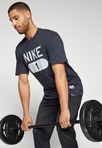 Marineblå Nike T-shirt med stort hvidt logo og nummer 1, parret med sorte træningsbukser. Model løfter en vægtstang.