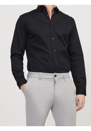 Homme portant une chemise noire boutonnée rentrée dans un pantalon gris clair sur mesure, les mains dans les poches, fond uni clair.