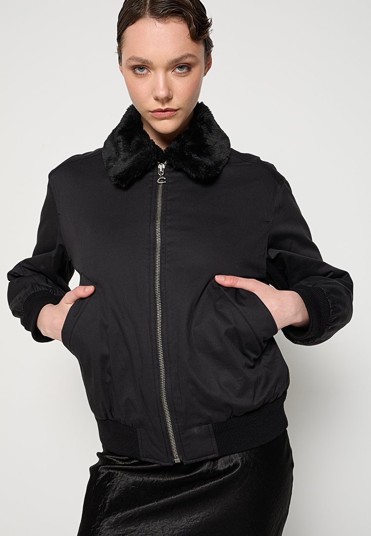 Czarna kurtka bomberka z sztucznym futrem przy kołnierzu, z zamkiem błyskawicznym z przodu i bocznymi kieszeniami. Materiał jest gładki z lekko błyszczącym wykończeniem.