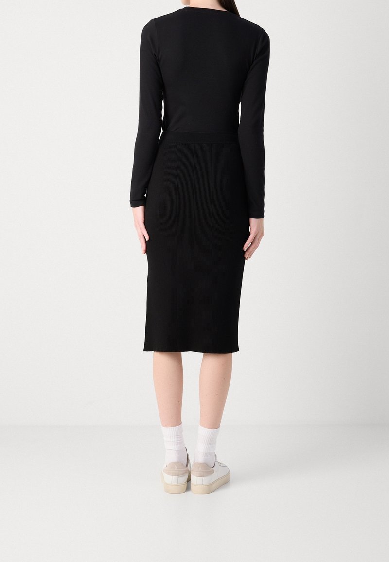 Femme portant un haut noir à manches longues et une jupe noire longueur genoux, chaussettes blanches et baskets blanches à plateformes, debout de dos.