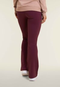 Leggings attillati e svasati in un profondo tessuto bordeaux con una consistenza liscia. Indossati con una maglietta beige chiara, abbinati a scarpe da ginnastica bianche.