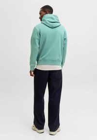 Minzgrüner Kapuzenpullover mit lockerem Sitz, gerippten Bündchen und einer Kängurutasche, kombiniert mit dunkelblauen, weit geschnittenen Hosen und hellen Turnschuhen.