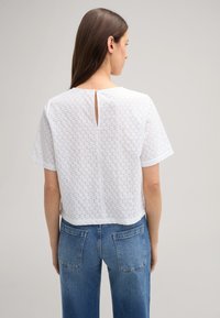 Witte top met korte mouwen en een bloemenpatroon, met een sleutelgat aan de achterkant en een textuurstof. Gecombineerd met blauwe high-waisted jeans met achterzakken.
