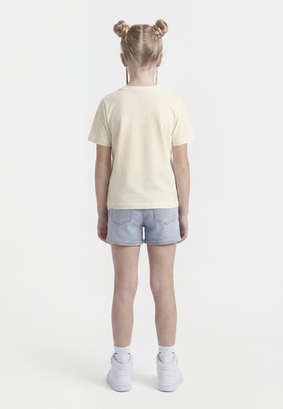 Otrok z blond lasmi v dveh figah, obrnjен stran, oblečen v kremasto majico, svetle denim kratke hlače, bele nogavice in bele superge.