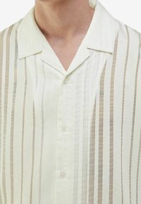 Vaalea nappikauluspaita, jossa on pehmeä tekstuuri ja vaaleanbeigeitä ja kermanvärisiä pystyraitoja. Klassinen kaulussuunnittelu pienillä napilla.