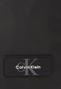 Étiquette noire Calvin Klein avec logo "CK" en relief. L'étiquette est rectangulaire avec des bords cousus et une texture mate.