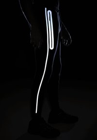 Melnas sporta bikses ar atstarojošiem akcentiem gar sānu šuvēm un rāvējslēdzēja kabatām, kas izceļ pieguļošu dizainu un gludu tekstūru.