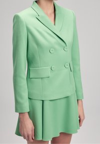 Zelený strukturovaný blejzer s dvojitými knoflíky, klopovým límcem a bočními kapsami. Sukně má rozšířený design, což vytváří celistvý outfit.