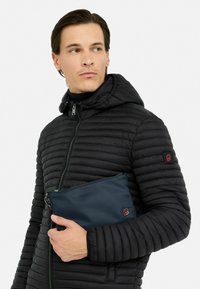 Čierna puffrová bunda s kapucňou, prešívaná štruktúra, predný zips a červený logový detail. Drží tmavomodrú kabelku.