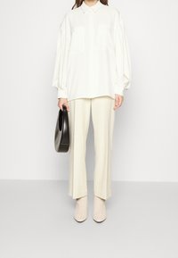 Camicia bianca oversize con bottoni e due tasche sul petto, abbinata a pantaloni crema a gamba larga e stivaletti beige a trama. Tiene una borsa nera.