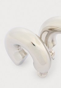 Cercei din metal cu un finisaj argintiu lustruit, având o formă curbată și abstractă. Designul este neted, fără embellishments vizibile.