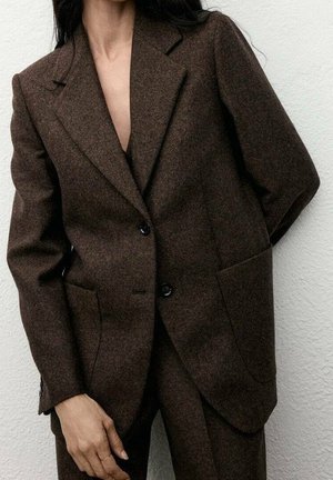 Femme portant un costume sur mesure marron foncé avec deux boutons, des revers crantés et des poches plaquées à l’avant, devant un mur clair uni.