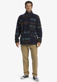 Fleece-Pullover in Dunkelblau mit mehrfarbigen geometrischen Mustern. Mit hohem Kragen und halbem Reißverschluss, kombiniert mit khakifarbenen Hosen und schwarzen Sneakers.