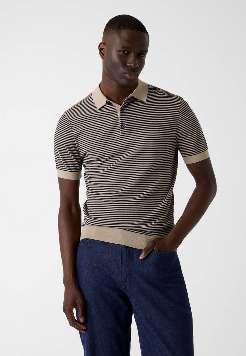 Un homme se tient debout, une main dans la poche d’un jean bleu, portant un polo à manches courtes, rayé beige et noir avec col et poignets beiges.