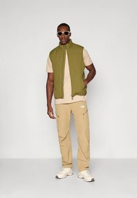 Alyvuogių žalios spalvos izoliuota liemenė dėvima ant smėlio spalvos marškinėlių ir smėlio spalvos darbo kelnių su užtrauktukais kišenėse. Dėvima su baltais sportbačiais ir akiniais nuo saulės.