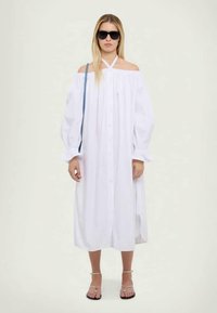 Bijela haljina bez naramenica s dugim rukavima, dugmadima sprijeda i bočnim prorezima, uparen s crnim naočalama i plavom torbom s remenom.