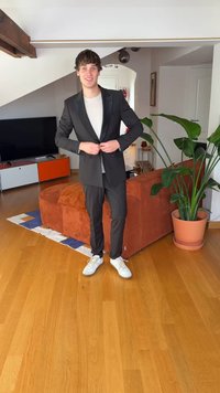 Sort jakkesæt med struktureret pasform, to-knaps lukning og notch revers, kombineret med hvide sneakers. Baggrunden viser en brun sofa og en plante.