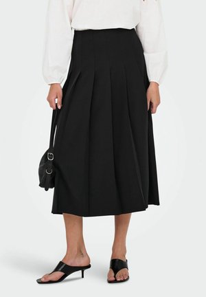 Woman wearing a black pleated midi skirt, white long-sleeve blouse, black heeled sandals, and holding a black handbag at her side.