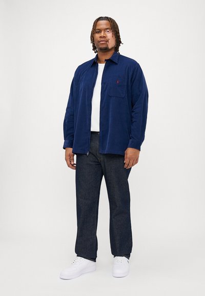 Μπλε κ�αμπαρντίνα μπλούζα με τσέπη στο στήθος, φορεμένη πάνω από ένα λευκό t-shirt και συνδυασμένη με σκούρα τζιν παντελόνια και λευκά αθλητικά παπούτσια.