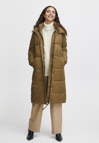 Duga maslinasto zelena puffer jakna sa patentnim zatvaračem, dva bočna džepa i kapuljačom. Nosi se preko kreme veste s pletenim uzorkom i bež hlača.