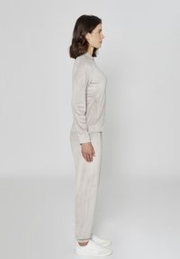 Jasnobeżowy dres wykonany z miękkiej tkaniny, z okrągłym dekoltem i bocznymi kieszeniami, zestawiony z białymi trampkami. Widok z profilu.