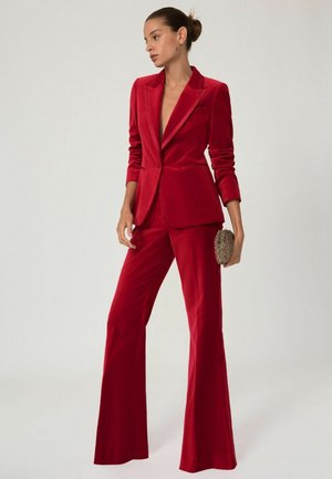 Abito in velluto rosso con giacca aderente e pantaloni svasati, caratterizzato da un profondo scollo e una clutch strutturata e decorata.