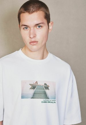 Young man with short hair wearing a white shirt featuring a dock leading to a boat on calm water at dawn.