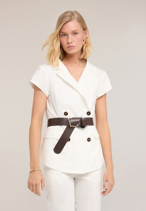 Lyshåret kvinde iført hvid kortærmet blazer med mørkebrun bælte og matchende hvide bukser, stående mod en neutral baggrund.
