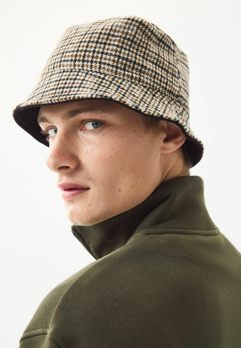 Bucket hat with a brown and beige houndstooth pattern, made from soft fabric. Model wears a green zip-up jacket with high collar.