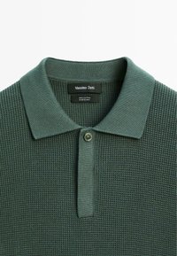 Polo in verde scuro, 100% cotone, tessuto a maglia testurizzato, colletto classico, pattina con un solo bottone, etichetta del marchio visibile sul colletto.