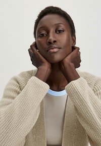 Cárdigan de punto texturizado beige con puños acanalados, llevado sobre una blusa ajustada de color claro con un cuello azul. Las manos descansan suavemente sobre el rostro del modelo.