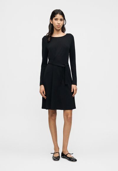 Robe noire à manches longues avec un corsage ajusté, une jupe évasée en forme de A et une ceinture nouée à la taille. Tissu à finition lisse et col rond.