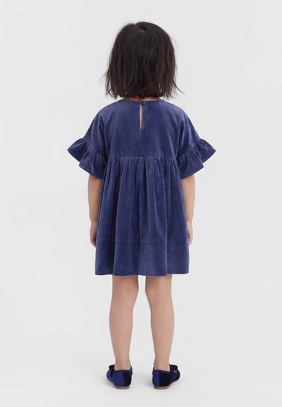 Barn med mørkt hår iført en løstsiddende, kortærmet blå fløjls kjole og matchende blå sko, vendt væk på en hvid baggrund.